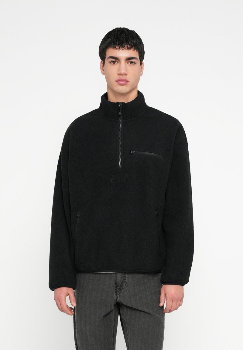 Νέος άνδρας με κοντά μαύρα μαλλιά, φοράει μαύρο φλις πουλόβερ με φερμουάρ και σκούρα γκρι τζιν, στέκεται μπροστά σε ένα απλό λευκό φόντο.