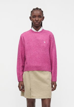 Persona con capelli intrecciati che indossa un maglione rosa sopra una camicia bianca e una gonna beige, in piedi su uno sfondo chiaro e uniforme.