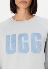 Light gray sweatshirt featuring a large, plush blue "UGG" logo. The fabric appears soft, with a crew neckline and raglan sleeves.