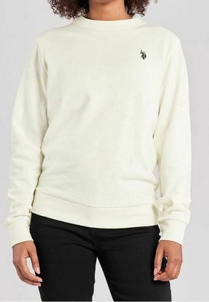 Personne portant un sweat-shirt blanc uni à manches longues avec un petit logo noir sur la poitrine, associé à un pantalon noir.