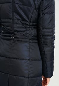 Svart vadderad jacka med quiltat mönster, som har dragskoaccentuer och metalliska detaljer i midjan. Slät, glänsande yta.