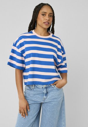 Femme aux cheveux tressés portant un chemisier à rayures horizontales bleues et roses et un jean bleu clair, debout avec une main dans la poche, sur un fond gris.