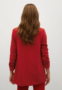 Blazer rojo a medida con mangas largas, un ajuste estructurado y una textura suave, visto desde atrás. Líneas limpias y sin elementos visibles de hardware.