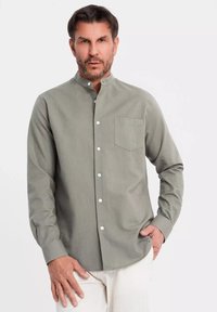 Camicia button-up verde realizzata in materiale morbido, caratterizzata da un colletto a banda, una tasca sul petto e bottoni bianchi, indossata con pantaloni chiari.