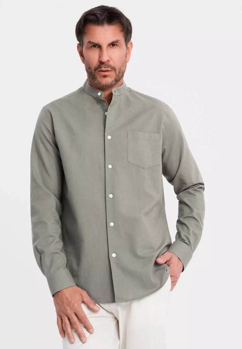 Camicia button-up verde realizzata in materiale morbido, caratterizzata da un colletto a banda, una tasca sul petto e bottoni bianchi, indossata con pantaloni chiari.