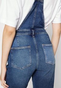 Jeans-Overalls in einem tiefblauen Farbton. Mit verstellbaren Trägern, Knopfverschluss und zwei Gesäßtaschen. Glatte Textur mit sichtbaren Nähten.
