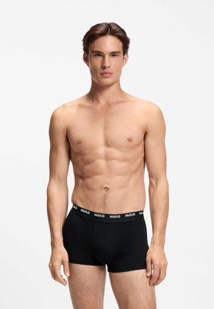 Młody mężczyzna z krótkimi ciemnymi włosami, ubrany w czarne bokserki Hugo, stoi na jednolitym białym tle.