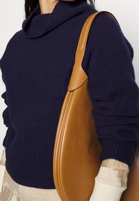 Pull côtelé bleu marine avec un col boule haut et des manches retroussées. Sac à épaule en cuir tan à texture lisse et au design incurvé.