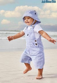 Salopette in cotone a righe azzurre, con bottoni e un piccolo dettaglio a tasca a forma di veliero, abbinata a un cappello a secchiello coordinato, indossata da un bambino sulla spiaggia.