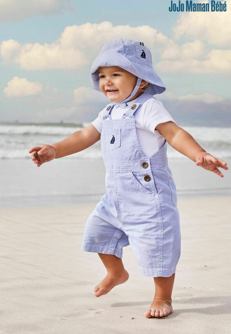 Salopette in cotone a righe azzurre, con bottoni e un piccolo dettaglio a tasca a forma di veliero, abbinata a un cappello a secchiello coordinato, indossata da un bambino sulla spiaggia.