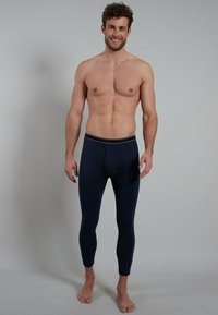 Smiling shirtless man standing barefoot wearing navy blue fitted long underwear pants against plain gray background.
