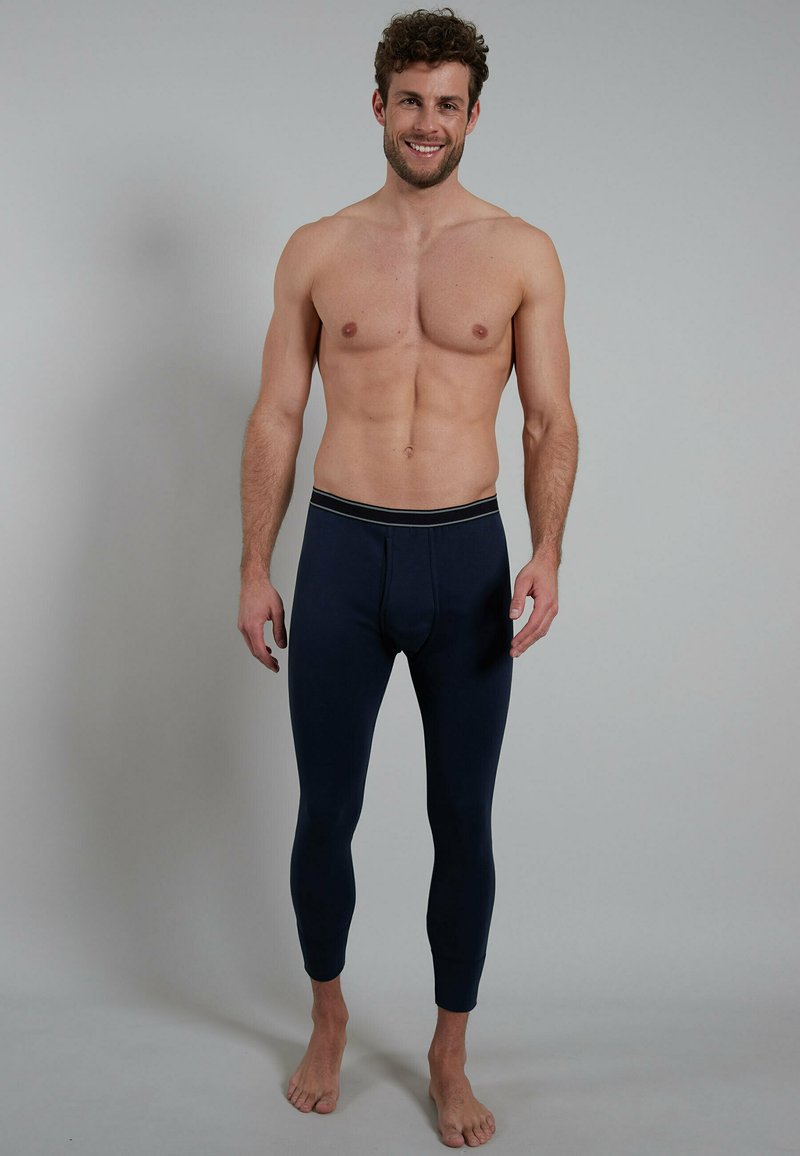 Smiling shirtless man standing barefoot wearing navy blue fitted long underwear pants against plain gray background.