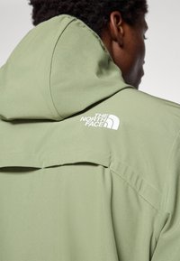 Person iført en lys grøn hættejakke med hvid "The North Face" logo på den øverste del af ryggen, vendt væk.