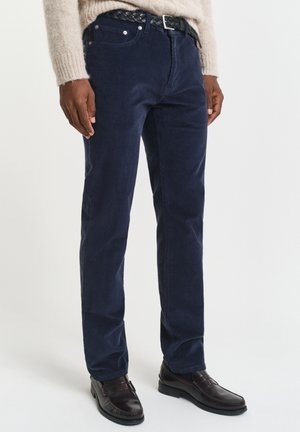 Mann med marineblå kordfløyelbukser og svart flettet belte, beige genser og mørkebrune skinnloafer, stående mot hvit bakgrunn.