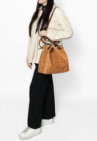 Tan leren emmerzak met een ingedrukt bloemmotief, bruine leren handvatten en een ronde bodem. Gedragen op de schouder met casual kleding.