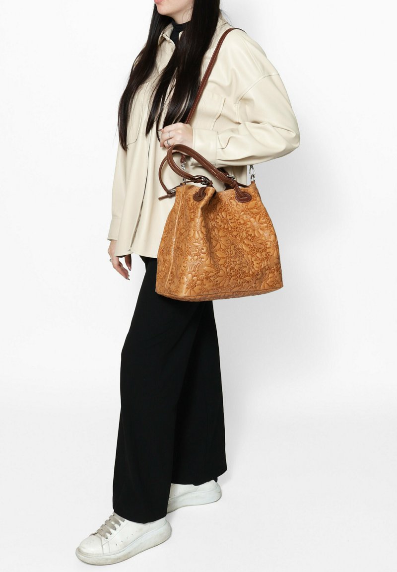 Tan leren emmerzak met een ingedrukt bloemmotief, bruine leren handvatten en een ronde bodem. Gedragen op de schouder met casual kleding.