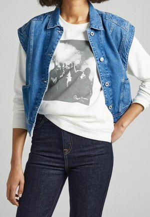 Veste sans manches en denim de couleur bleue contrastante, à la coupe ajustée, avec des boutons en métal, superposée à un sweatshirt blanc avec un imprimé graphique noir.