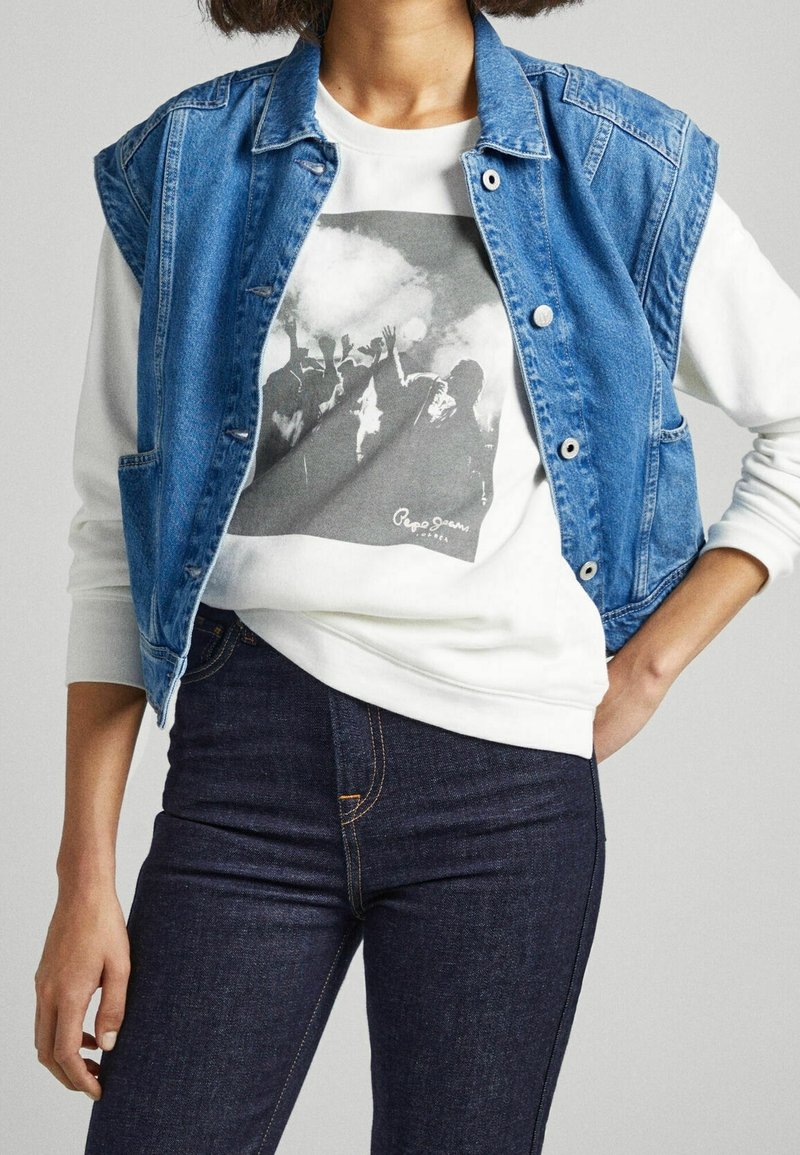Giacca in denim senza maniche di colore blu a contrasto, dalla forma aderente e con bottoni metallici, indossata sopra una felpa bianca con stampa grafica nera.