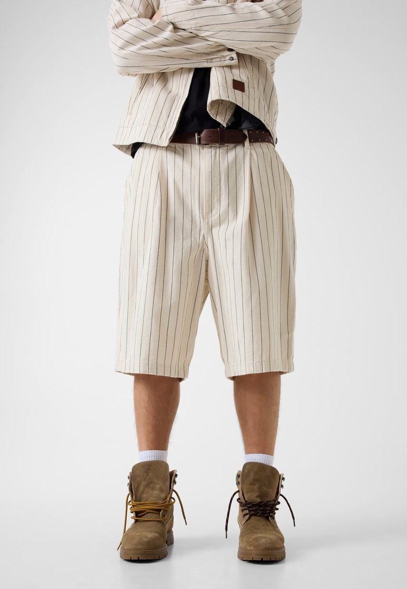 Personne portant un short beige à fines rayures et une veste assortie, ceinture marron, chaussettes blanches, et bottines marron dépareillées à lacets, debout les bras croisés.