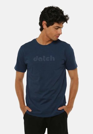 Uomo con una maglietta blu navy con il logo "datch", mani in tasca, guarda verso il basso a sinistra, su uno sfondo chiaro e uniforme.