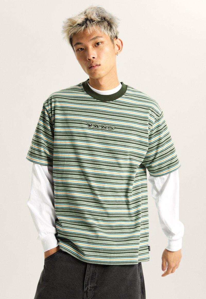 Grønstribet t-shirt med marineblå, orange og hvide linjer. Har en rund halsudskæring og logodetalje i midten. Bæres over en hvid langærmet skjorte.
