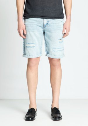 Uomo che indossa pantaloncini di jeans azzurri strappati, scarpe nere e una camicia nera, in piedi davanti a uno sfondo bianco semplice.