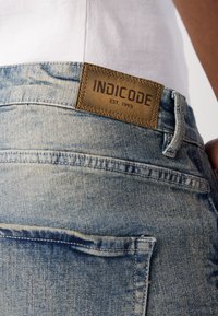 Denim jeans in light blue, featuring a brown leather label with "INDICODE EST. 1993." Visible stitching and a back pocket detail.