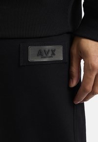 Czarne odzież sportowa z prążkowaną teksturą i wyraźną metką z literami "AVX." Zbliżenie na materiał i ułożenie ręki.