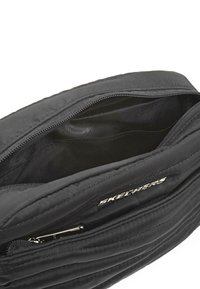 Bolso acolchado negro de Skechers con cremallera principal abierta que muestra un interior vacío y un bolsillo frontal adicional con cremallera.