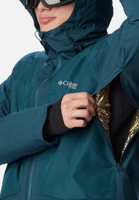 Türkise Outdoor-Jacke mit Kapuze, diagonalem Reißverschluss, schwarzem Innenfutter und goldfarbener reflektierender Beschichtung in der Tasche.