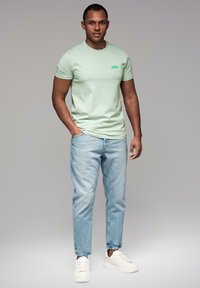 T-shirt verde menta a girocollo con un piccolo logo a contrasto, abbinata a jeans azzurri e sneakers bianche, in piedi davanti a uno sfondo neutro.