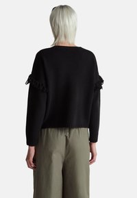 Černý zkrácený svetr s texturovaným třásněním na rukávech, pletená látka a žebrované manžety. V kombinaci se světle zelenými, volnějšími kalhotami.