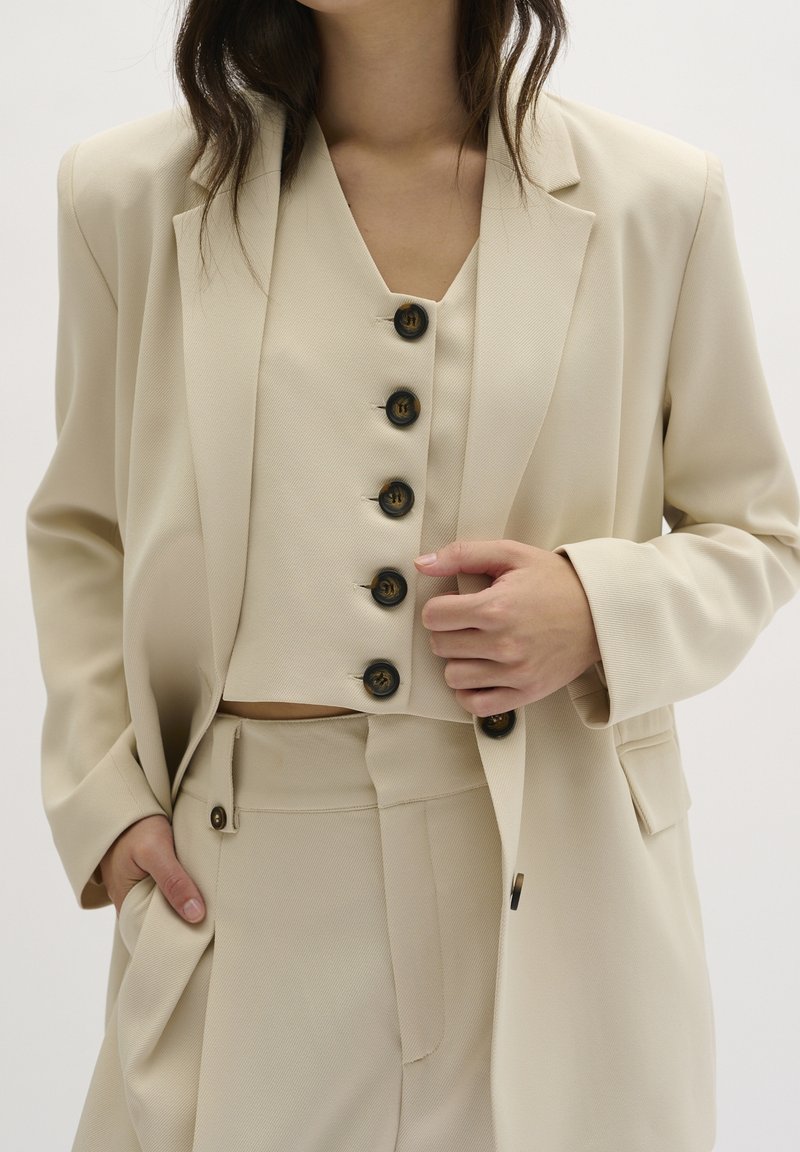 Blazer beige ajustado sobre un chaleco a juego con seis botones oscuros y pantalones de pierna ancha. Tejido texturado con un acabado suave.