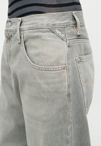 Ανοιχτό γκρι τζιν παντελόνι με τσέπες μπροστά και πίσω, χάλκινα καρφιά και ετικέτα «Replay» πάνω από την μπροστινή τσέπη, φορεμένο με μαύρο ριγέ τοπ.