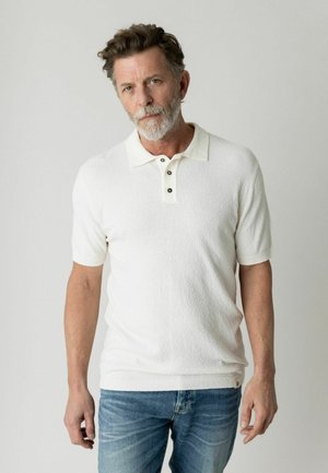 Mittelalter Mann mit grauem Bart, der ein weißes kurzärmeliges Poloshirt und blaue Jeans trägt, vor einem schlichten hellen Hintergrund stehend.