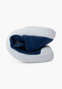 Scarpa slip-on di tela blu con suola in gomma bianca, caratterizzata da una forma arrotondata e una texture di tessuto morbido, piegata all'interno per mostrare l'interno.