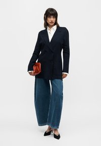 Woman wearing black double-breasted blazer, white shirt, wide-leg blue jeans, black heels, holding folded red clutch bag, standing against white background.