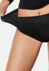 Gros plan d'une personne en train d'étirer la ceinture d'un sous-vêtement noir, montrant son élasticité et son ajustement autour des hanches et de la taille.