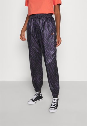 Personne portant un pantalon de jogging Nike noir à imprimé zèbre violet avec des poignets élastiques, associé à des baskets montantes Converse noires et un t-shirt orange.