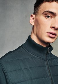 Veste zippée vert foncé avec col haut, comportant des panneaux matelassés et une texture lisse. Zip en métal et détails de couture minimaux.