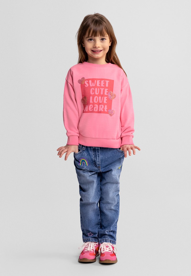 Fille souriante portant un sweat-shirt rose avec le texte "Sweet Cute Love Heart", un jean bleu avec un patch arc-en-ciel, et des baskets roses, debout sur un fond gris.