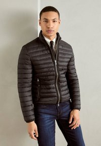 Veste noire rembourrée avec des coutures horizontales, col haut et fermeture éclair à l'avant. Elle présente deux poches latérales et une coupe ajustée, associée à un jean foncé.