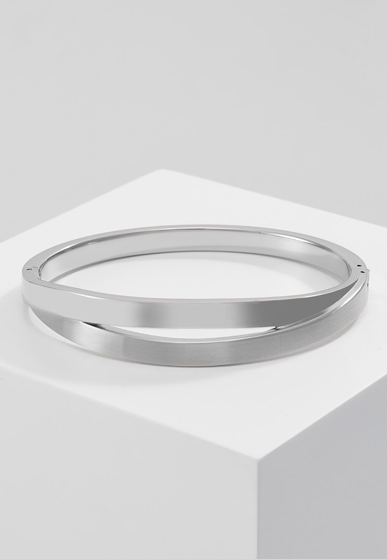 Bracelet jonc en métal argenté lisse avec un design épuré et plat, reposant sur un cube blanc sur un fond gris uni.