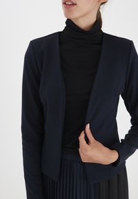 Tmavě modrý elastický svetr s dlouhými rukávy a otevřeným předním dílem, vrstvený přes černý rolák. Ruka umístěná na okraji svetru.