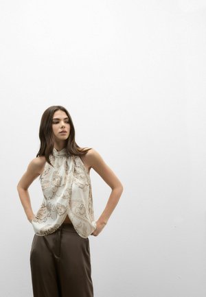 Jeune femme aux longs cheveux bruns portant un haut blanc sans manches à motif cachemire et un pantalon marron, debout, mains sur les hanches, contre un mur blanc uni.