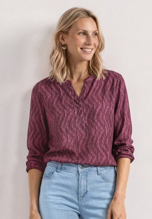 Uśmiechnięta kobieta o blond włosach do ramion, ubrana w bordową, teksturowaną koszulę z długim rękawem i jasnoniebieskie dżinsy na tle jednolitej ściany.