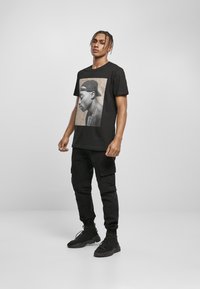 Schwarzes Baumwoll-T-Shirt mit einem monochromen Grafikdesign eines männlichen Körpers, getragen mit schwarzen Cargo-Hosen und schwarzem Schuhwerk, vor einem neutralen Hintergrund.