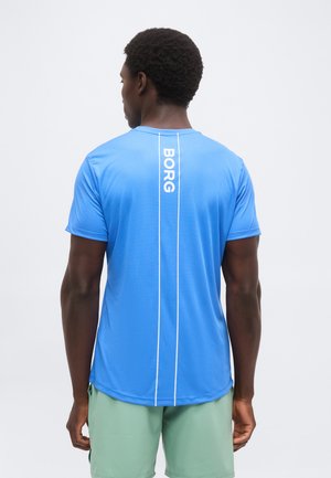 Homme vu de dos portant un t-shirt sportif bleu à manches courtes avec le texte « BORG » dans le dos et un short vert clair sur fond uni.