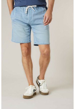 Homme portant un short en jean bleu clair avec un cordon blanc, une chemise bleu marine et des baskets blanches à rayures noires.