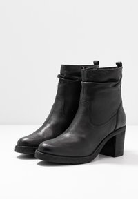 Botas de tobillo de cuero negro con un diseño ligeramente arrugado, tacón texturizado y un tacón alto y grueso. Cuentan con una cremallera lateral para facilitar su uso.